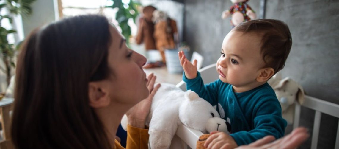 Cute baby boy in pajamas trying to explain something to his mother while standing in crib early in the morning