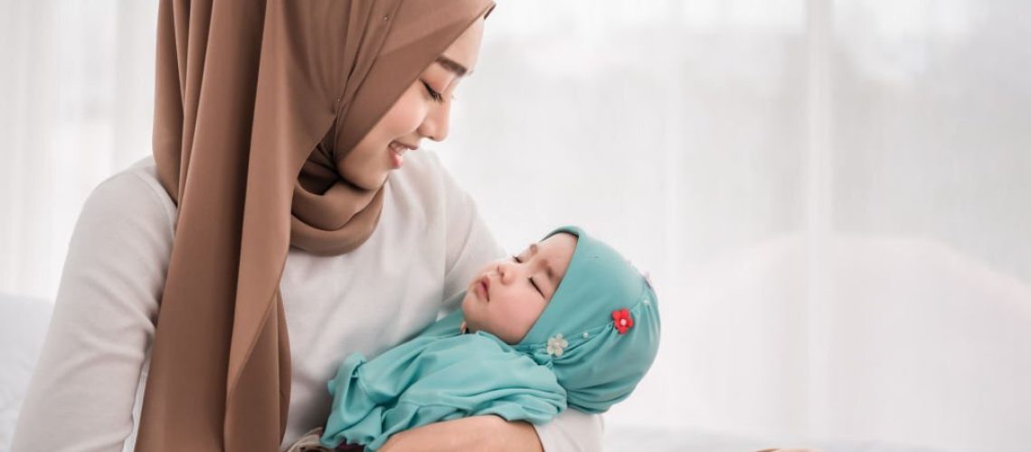 Happy muslim mother holding adorable little baby daughter wearing hijab in her arms on white bed in bedroom. Arab young mom wearing a head scarf, and she's sitting on the bed while holding her baby.