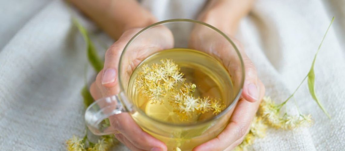 A cup of herbal tea (lime blossom/ linden tea) photographed on a cozy beige blanket with herbal flowers in the background.