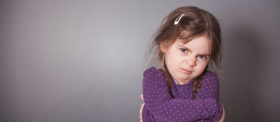 A little cute Girl is sulking. She is wearing a purple shirt in front of a gray background.