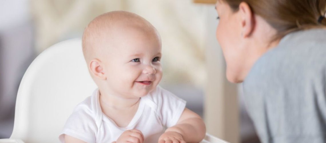 An adorable baby sits in a high chair and smiles at her mother as she reaches for finger food on the tray.