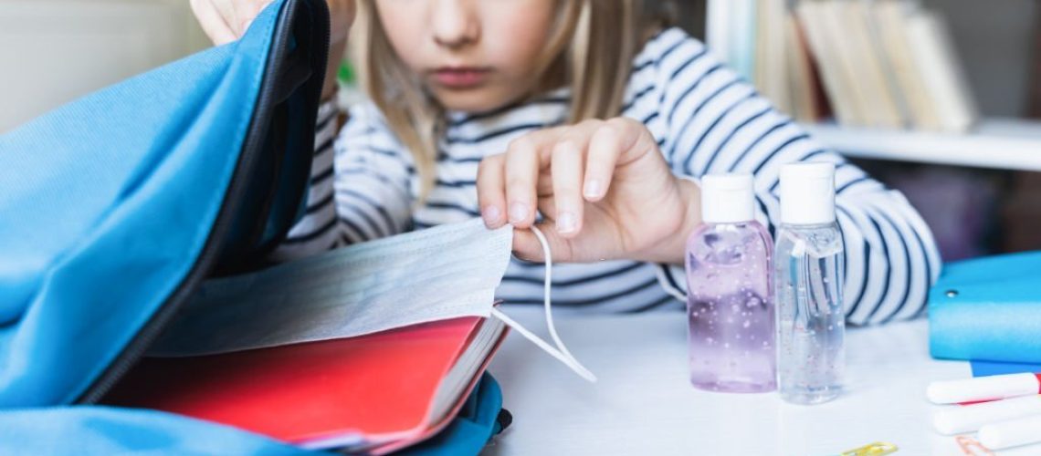 Little girl packing blue backpack in kids's room. Face mask, bottle of sanitizer, stationery, pens, multicolored markers, notebooks. Back to school. Mom's hygiene,safety precautions after coronavirus.